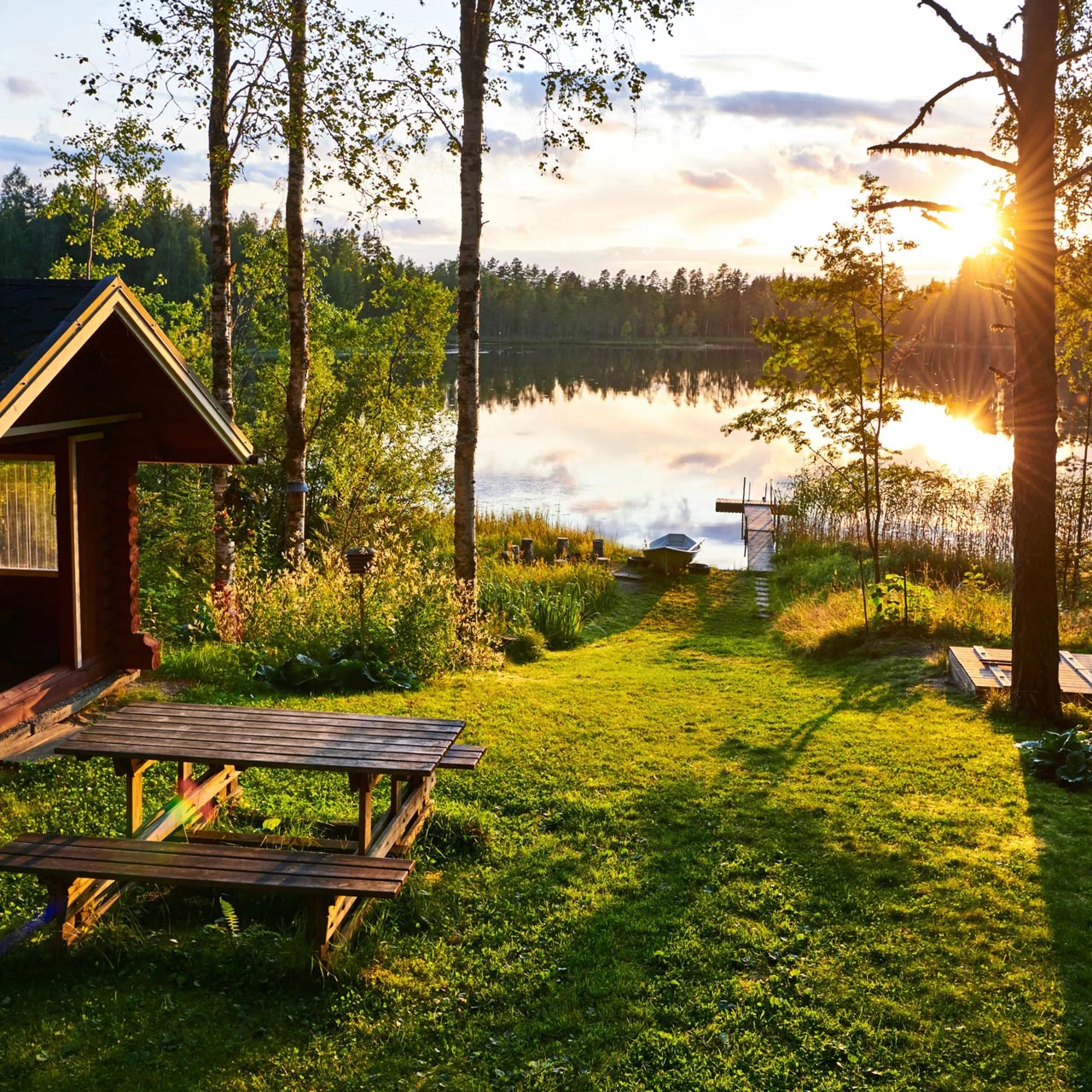 Finnisches Sommerhaus aus Holz am Seeufer bei Sonnenuntergang – idyllische Landschaft mit grüner Wiese, Bootssteg und spiegelglattem Wasser, umgeben von Bäumen und Natur