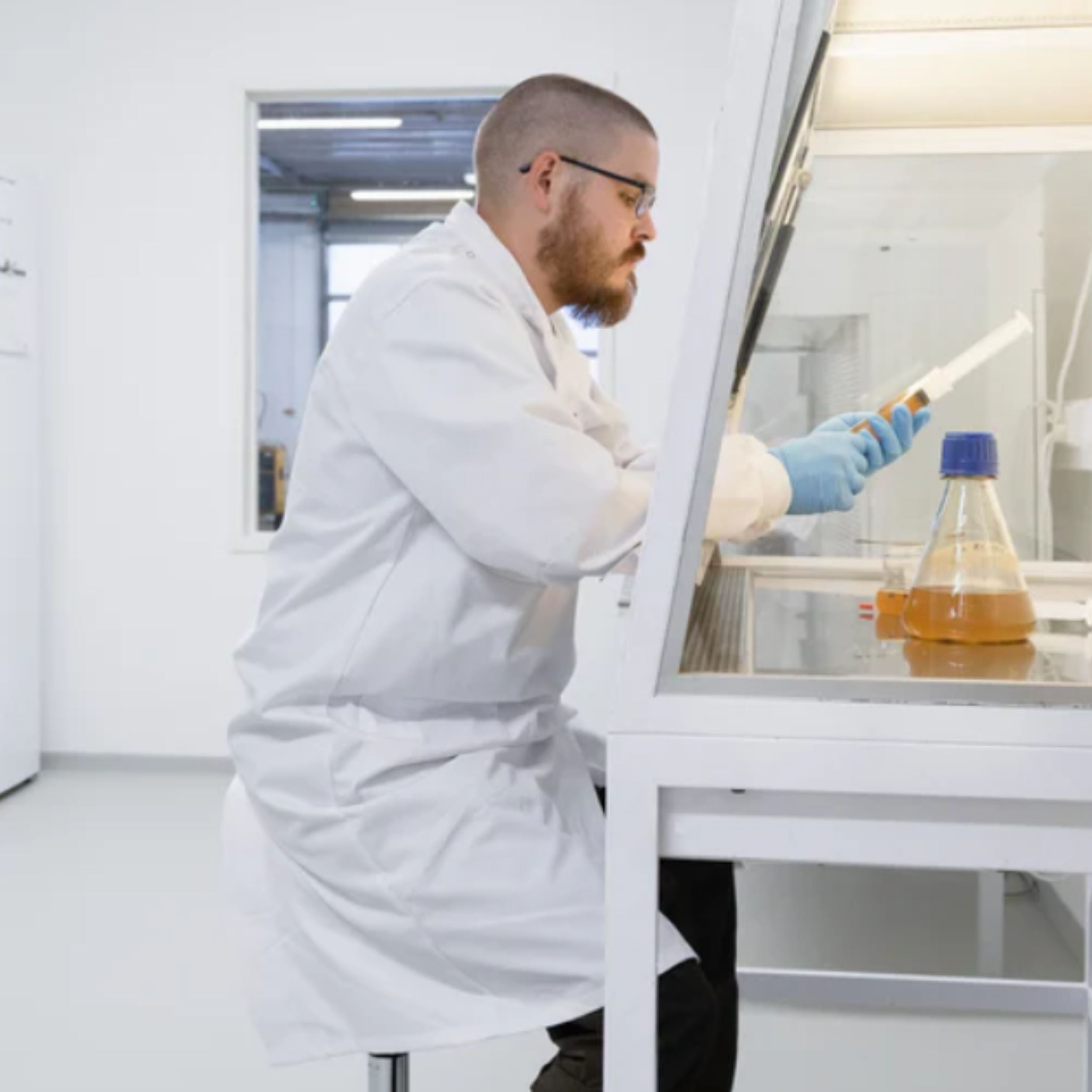Laborant in weißem Laborkittel arbeitet mit einer Spritze an einer Arbeitsstation, daneben ein Erlenmeyerkolben mit orangefarbener Flüssigkeit. Das Bild symbolisiert die sorgfältige wissenschaftliche Arbeit und Qualitätskontrolle bei der Herstellung von Pilzextrakten.