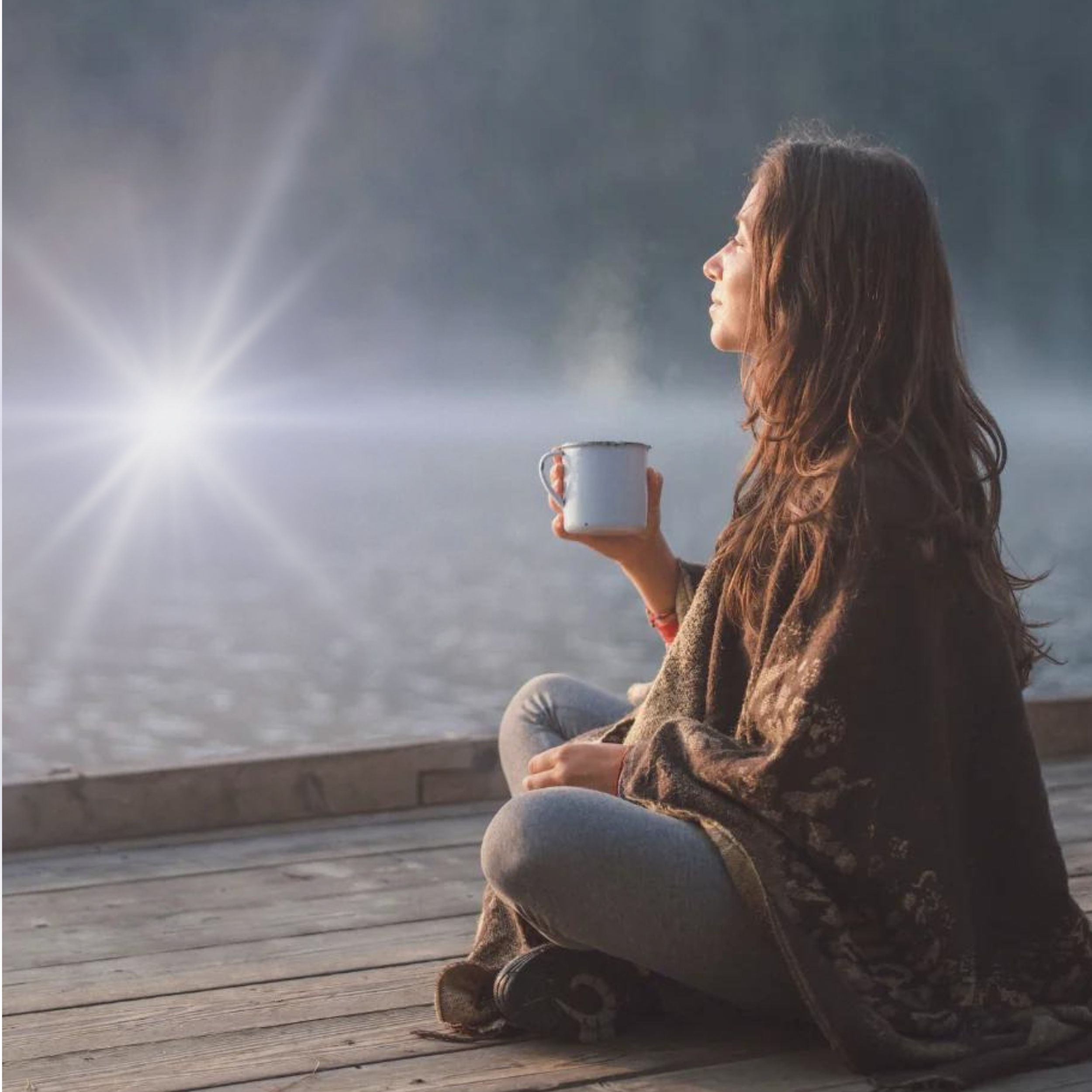 Frau sitzt in eine Decke gehüllt auf einem Steg am See und hält eine dampfende Tasse in der Hand, während die Sonne über dem Wasser glitzert – Moment der Ruhe und Entspannung in der Natur.